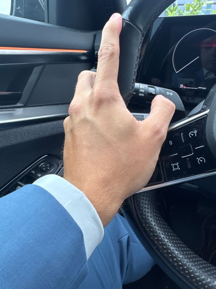 Eine Hand am Lenkrad eines Autos, man sieht einen blauen Anzug und ein Hemd