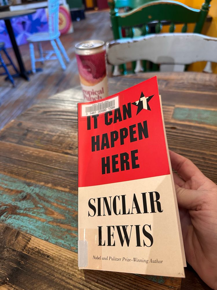 Picture of book “It can’t happen here”, by Sinclair Lewis published in 1935. To the right of the book is a can of kombucha that sits on a distress finished oaken table in an organic vegan cafe in Lowell, MA.