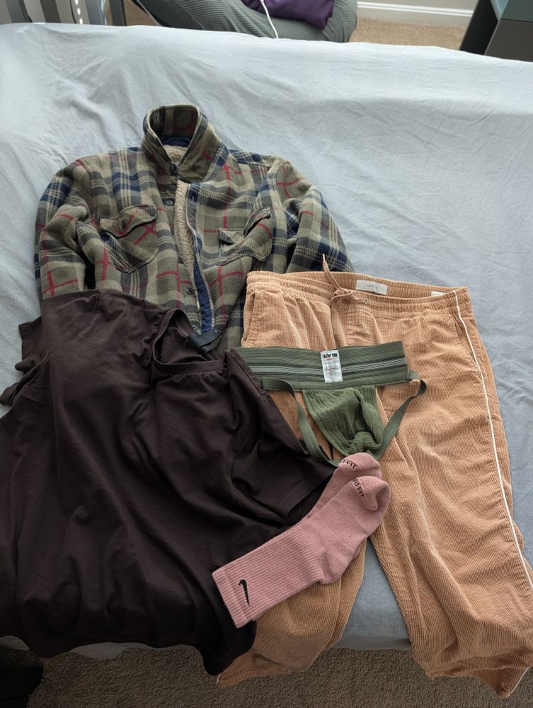 Clothes layered on top of each other, olive green fleece jacket, tan joggers, brown crew neck t shirt, pink socks, and an olive green jockstrap on a grey bedsheet