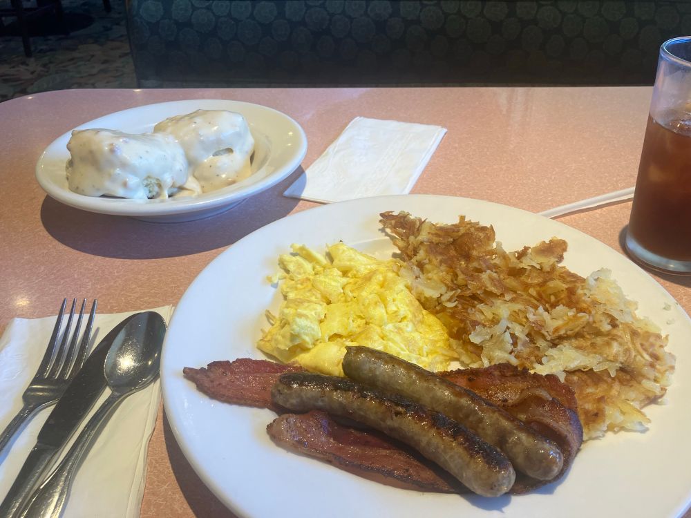A breakfast of scrambled eggs, 2 link sausage, two bacon slices, hash browns, and a pair of biscuits with gravy on a separate plate. A glass of iced tea, along with utensils, are nearby.