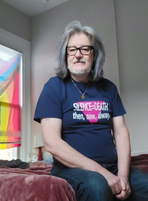 A 60-something white queer male is sitting on a burgundy coloured blanket on the side of a bed. They are wearing a dark blue t-shirt with "SILENCE=DEATH then, now, always" in white text across a vibrant pink triangle. They are wearing jeans, and their hands fall gently in between their knees. Their hair is mostly grey. They're wearing dark framed prescription glasses and have a small gold coloured necklace. Over their right shoulder a part of a Progress Pride flag is visible.
