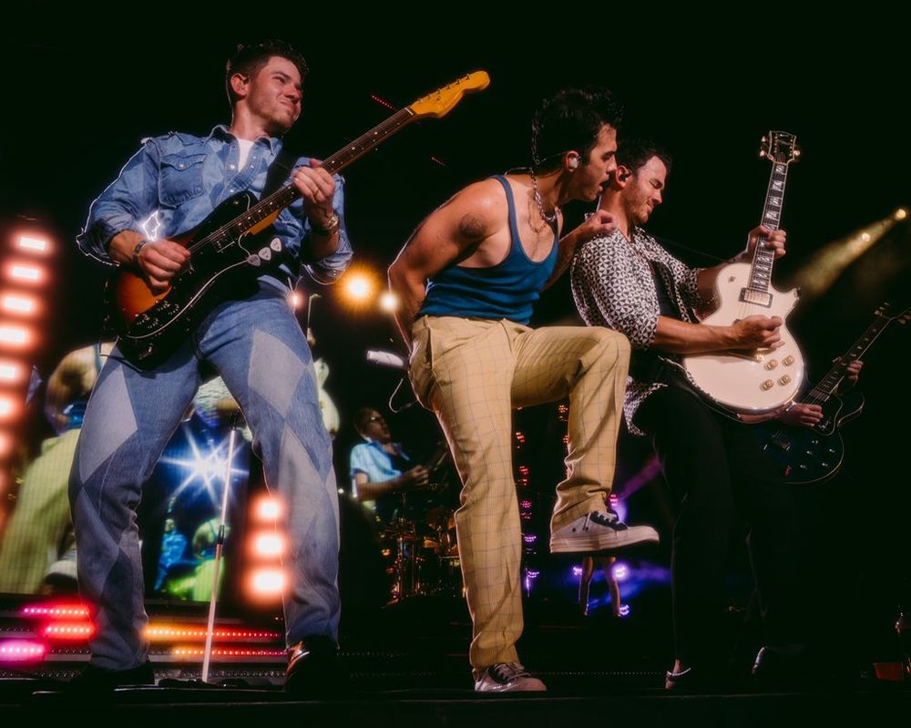 Jonas Brothers in concert. Nick and Kevin with guitars and very expressive faces. Joe in the middle with one knee up.