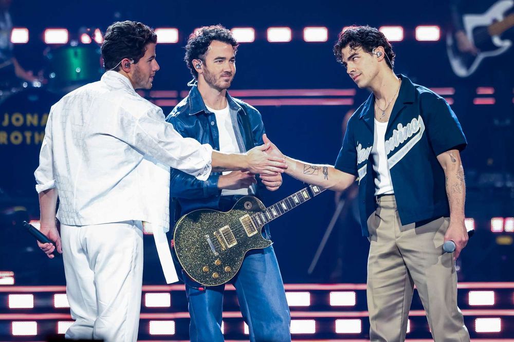Jonas Brothers in concert. Nick in white, Kevin in full jean and Joe wearing a baseball shirt. Nick and Joe are looking at each other and shaking hands. Kevin looks at the crowd with a grateful expression.