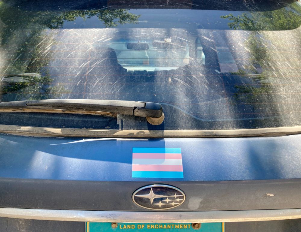 The back of a blue 2007 Subaru Legacy Outback with a very dirty rear window and a transgender pride flag above the Subaru logo.