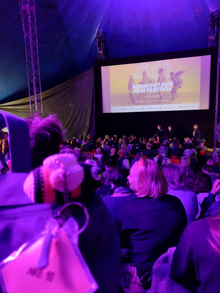 Movie audience of few hundred people in a circus tent, with a screen with the words "Midnight Sun Film Festival" on a yellow background and a small crocheted bee plushie on the foreground.