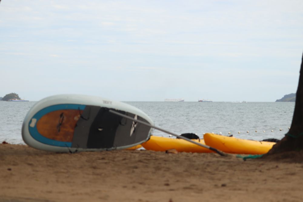 Uma prancha de stand-up sobre a areia, apoiado no remo e dois caiaques amarelos atrás, também sobre a areia