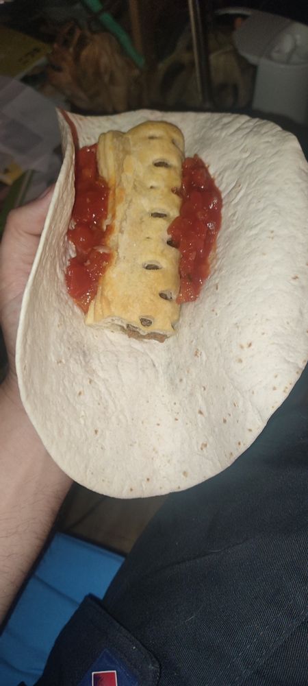 A white flour tortilla sits open in a white guy's hand. In the centre of the tortilla is a golden brown sausage roll flanked by two dollops of salsa
