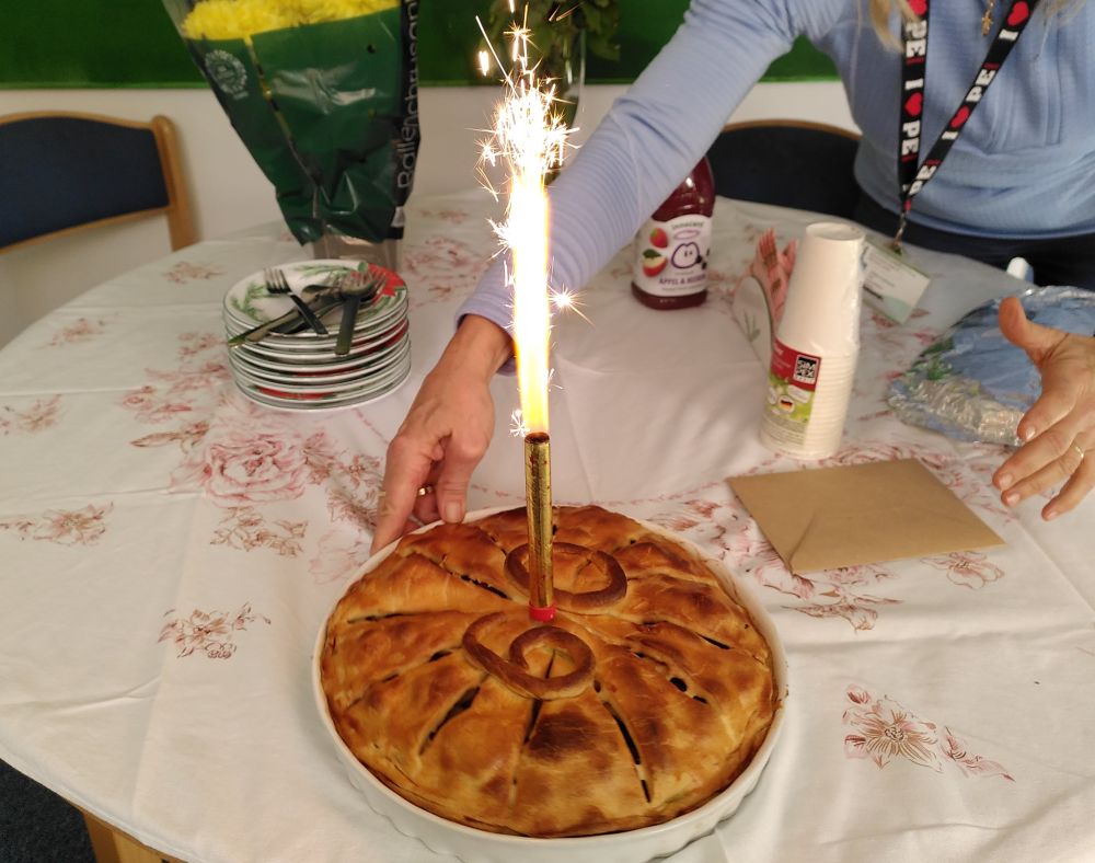 Apple pie in white dish with "60" on top crust and sparkler fired up in the middle; colleague's hands are positioning the pie on a round table with table cloth.