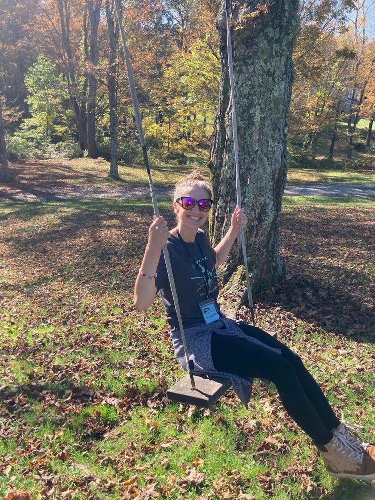 Photo of Kiley on a tree swing. She is a white woman with hair in a bun, smiling and wearing sunglasses.