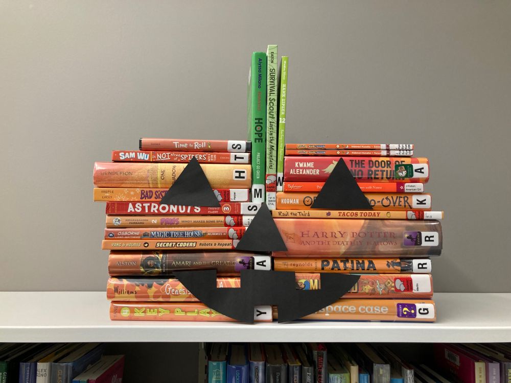 Middle grade novels and graphic novels with orange spines stacked to look like a pumpkin. Black cardboard cutouts are taped to the front to make the stack look like a Jack-o-lantern. Three books with green spines are stacked vertically to resemble a stem.