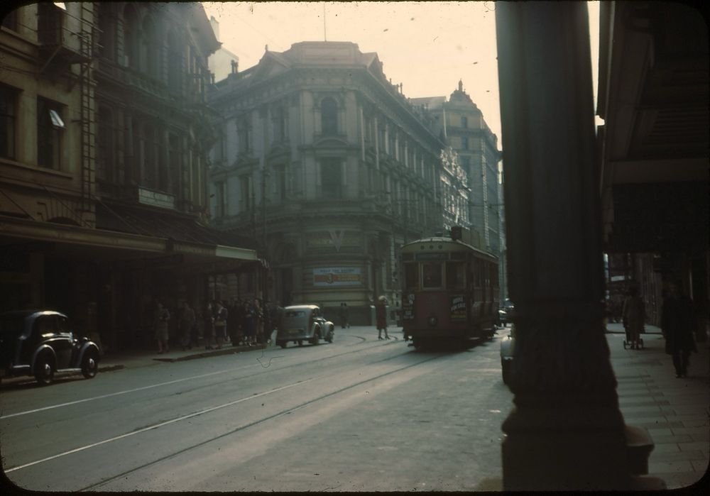 On a slightly overcast winter’s day in the central business district of Wellington, a “double saloon” tram has cleared the junction of Lambton Quay and Willis Street as pedestrians begin to cross behind it. The tram’s destination is just visible: it’s on route 2, an inner-city shuttle service, running to Courteney Place from Wellington Railway Station, with advertising signs for Lux soap and a real estate sale on its front cab. This photo was taken exactly a year after the Pacific arena of the Second World War had come to an end and, to mark the anniversary, the Bank of New Zealand building behind the tram has a letter “V” above its curved corner billboard 