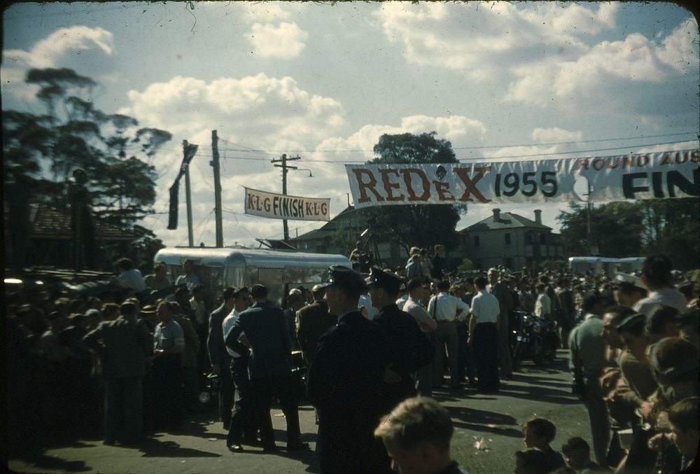 Looking towards the Redex Round Australia Reliability Trial 1955 road race finish line, marked by banners over a street in Western Sydney. Among the crowd of spectators are a pair of police officers, reporters, photographers, newsreel camera operators on top of vans and several steel caravans 