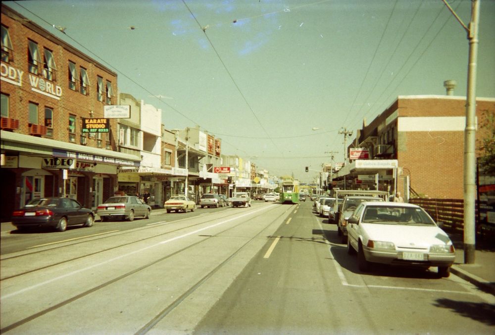 A slightly faded and stained photograph looking along a typical inner suburban Australian Main Street. In the middle distance an angular nineteen-seventies tram, in its eighties-into-nineties green and yellow colour scheme, is heading towards the camera