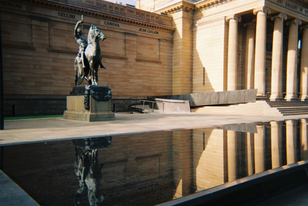 On a sunny, slightly smoky morning, a bronze equestrian statue, standing in front of a yellow-brown classical revival sandstone building, is reflected in the still water of a simple ornamental pool