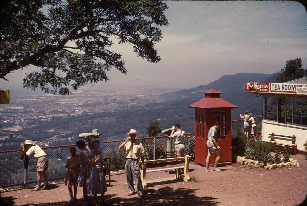 Looking along an escarpment above a large regional city towards a group of sightseers at a lookout, taking photos, looking through telescopes or just walking around. The clothes suggest a warm day: one man, slightly more formally dressed than everyone else, is drinking from a glass soft drink bottle, presumably supplied by the local Parkinson’s company, as advertised by the tea room at right, just past a rather Tardis-shaped red telephone booth