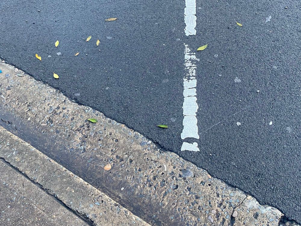 A close up view of an angled parking space, with the white line fragmented across the asphalt, the section leading to the concrete gutter vaguely resembling an exclamation mark 