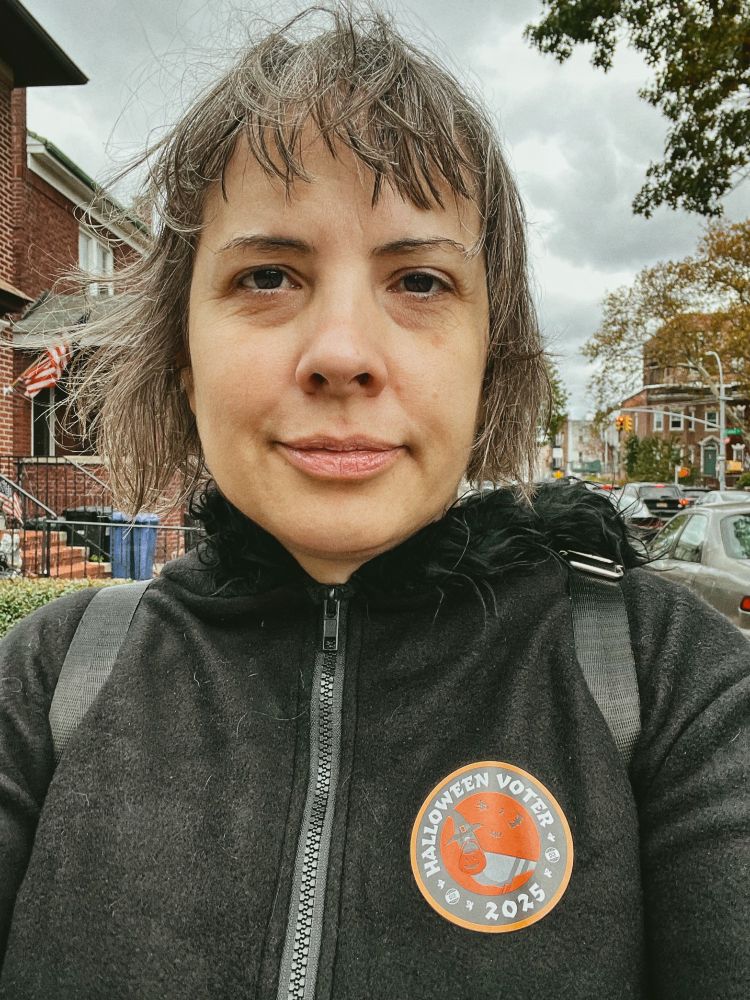 Selfie of me walking down the street with a slight smile, a Halloween Voter 2025 sticker affixed on my fleece hoodie