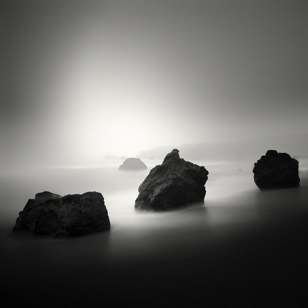 This is an image of the sun piercing through a misty day along the Sonoma Coast of Northern California on the West Coast of the United States. In the foreground are three rocks and dark sand. In the distance are cliffs shrouded in the mists. 