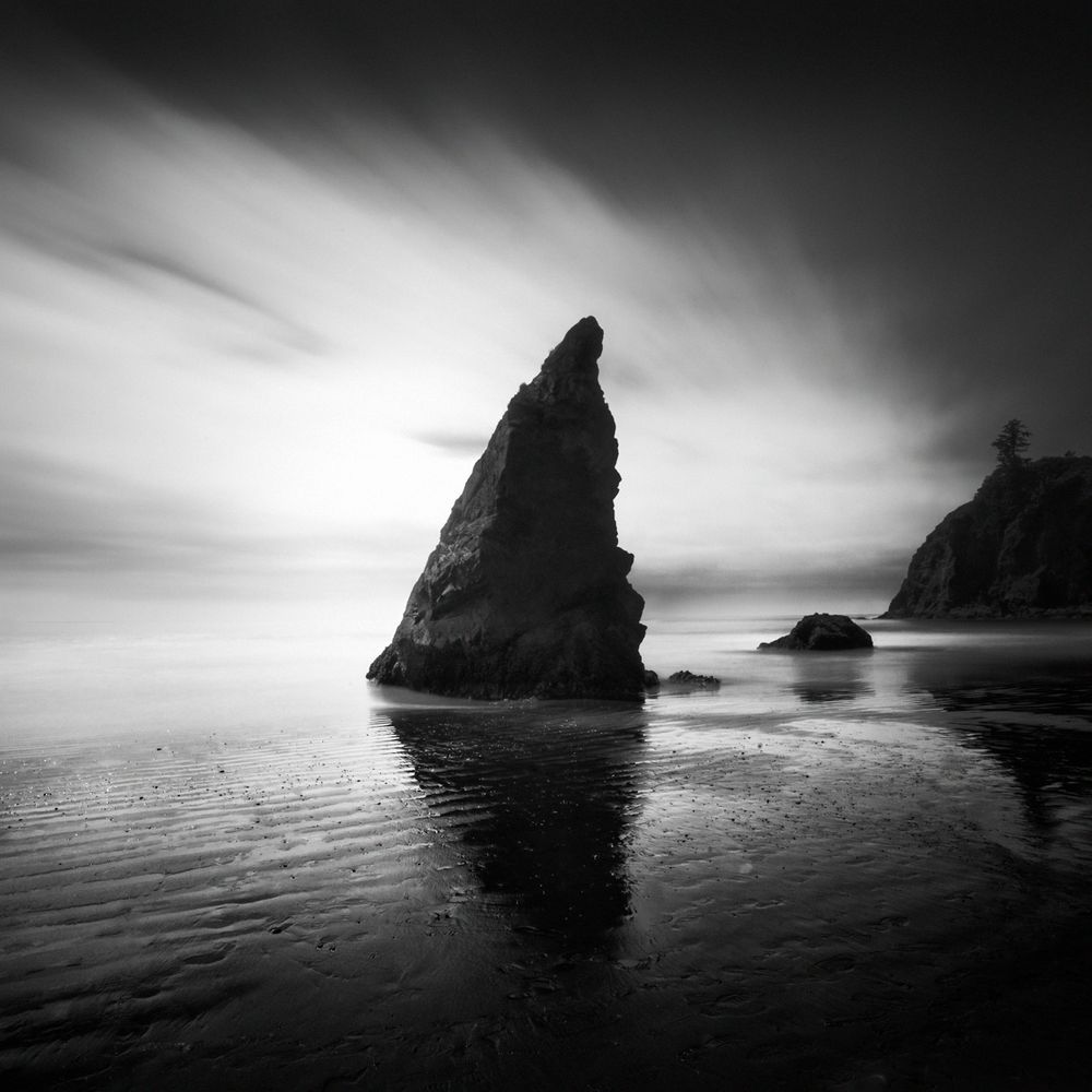 This is a black and white image of a rectangular rock on the shoreline-- with part of an island-like sea stack in the distance on the right. The wet sand includes reflections of the sea stacks. 