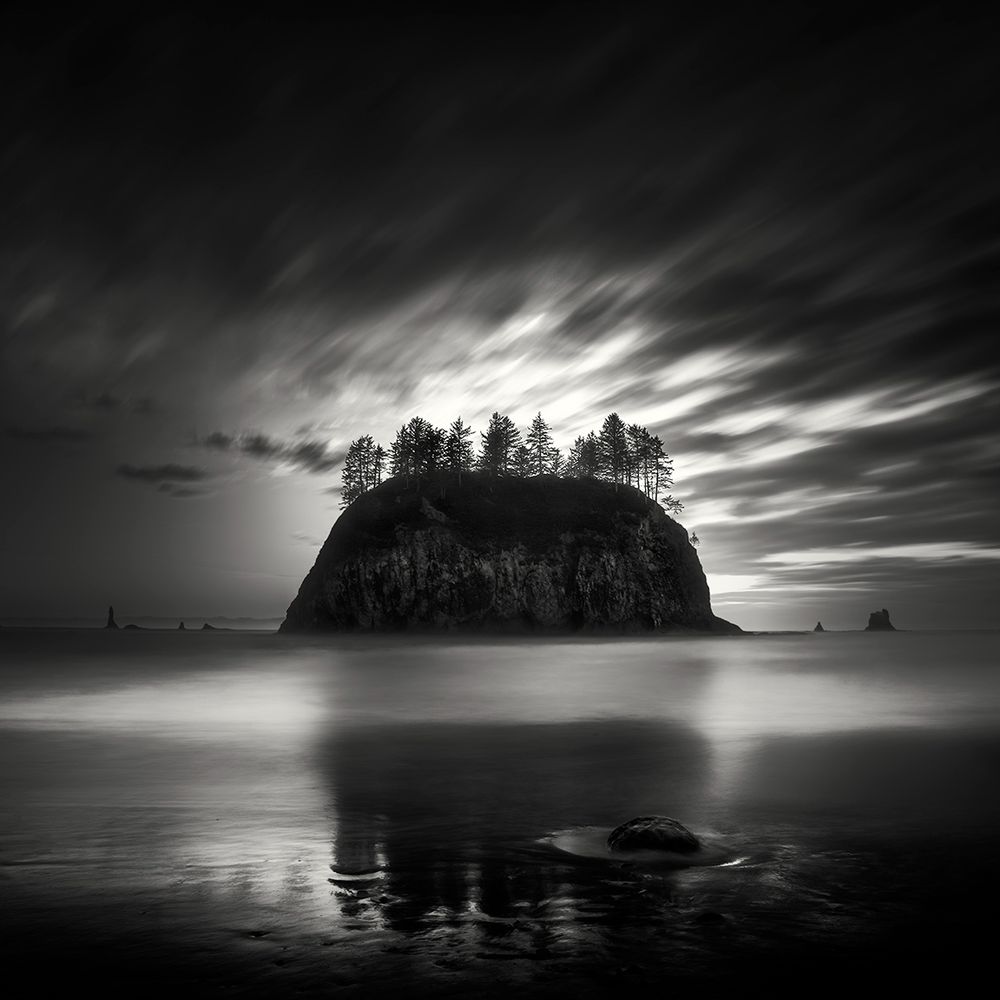 This simple, basic photograph is a black and white image of an island-like sea stack sitting in the water just off the shoreline.  A gathering of trees grows from its base. The sea stack's reflection can be seen in the wet sand.  The sun shines in the background-- radiating above the sea stack. 