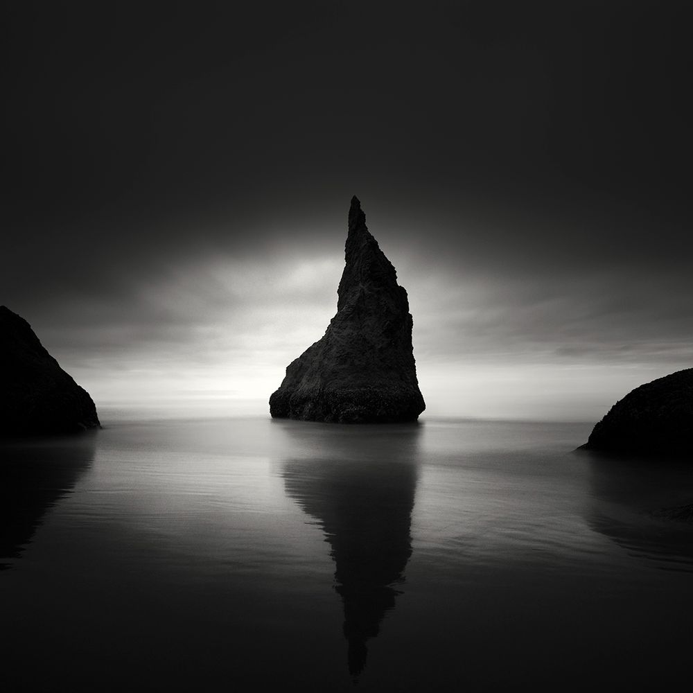 This is a picture of a rock that looks a lot like a witch or wizard hat. It sits on the shoreline with light and clouds in the background.   