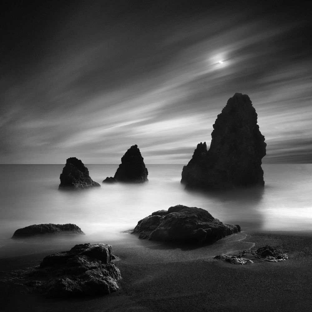 This is a black and white image of a trio of rocks in the water and four rocks in the sand and water of the shoreline. In the distance, the light breaks through the clouds. 