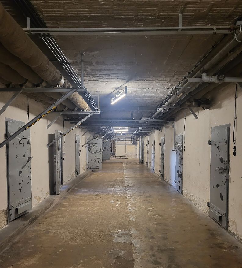 Photo d'un long couloir au sol brun sale, avec des murs blancs des 2 côtés avec des portes grises de prison. Au plafond courent de longs tuyaux et se trouvent des néons qui donnent un éclairage très cru.  