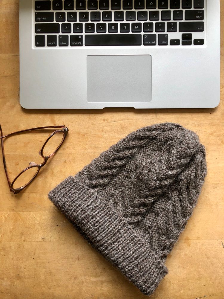 A flat lay photo of a light brown cabled toque against a wood tabletop with a pair of glasses and a laptop keyboard visible. 