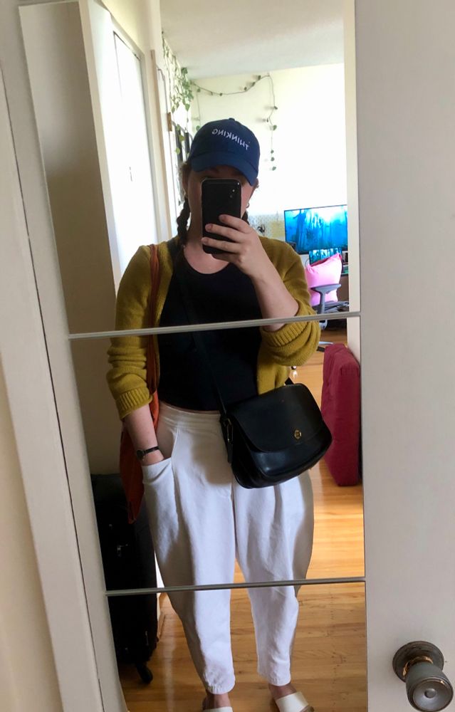 I take a mirror selfie wearing me-made white barrel leg pants, a black crop top, and thrifted pickle green sweater. I have a black leather vintage coach bag worn across my body and an orange tote bag filled with library books on one shoulder. To top it off is my blue thinking cap 🧢😜