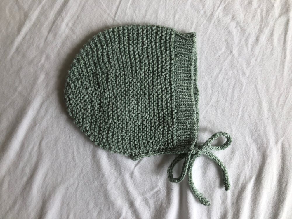 A flat lay photo of a seafoam green-blue baby bonnet knit in garter stitch with ribbed brim and I-cord ties.
Pattern is Bearly Bonnet - Pure Stitches (mods: no ears, added i-cord finish and ties)