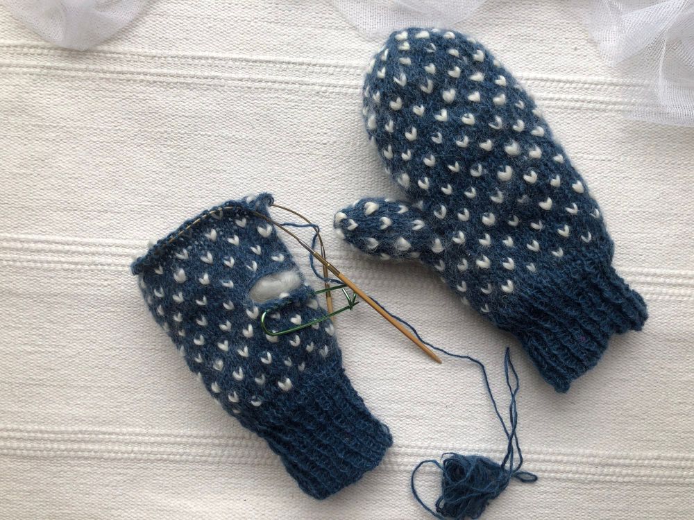 A flat lay photo showing one complete blue handknit mitten with natural roving thrums and one mitten in progress.