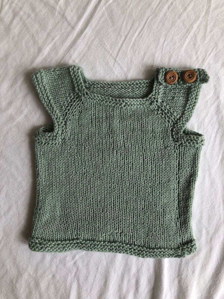 A flatlay photo of a seafoam green-blue newborn vest with light wooden buttons at one shoulder, knit in stockinette stitch with garter stitch hems.
Pattern is Baby T-Shirt Vest - Sam Lamb