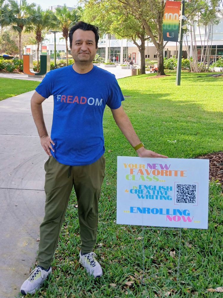 A good-looking South Asian man wearing a blue t-shirt that reads "FREADOM" stands next to a sign promoting the English & Creative Writing Department at the University of Miami, with the university's signature "U" sculpture visible behind his right shoulder and a banner celebrating the university's centenary in the right background. The sign reads "Your New Favorite Class // English & Creative Writing / Enrolling Now" with a QR code that links to the course descriptions of next semester's ENG classes.