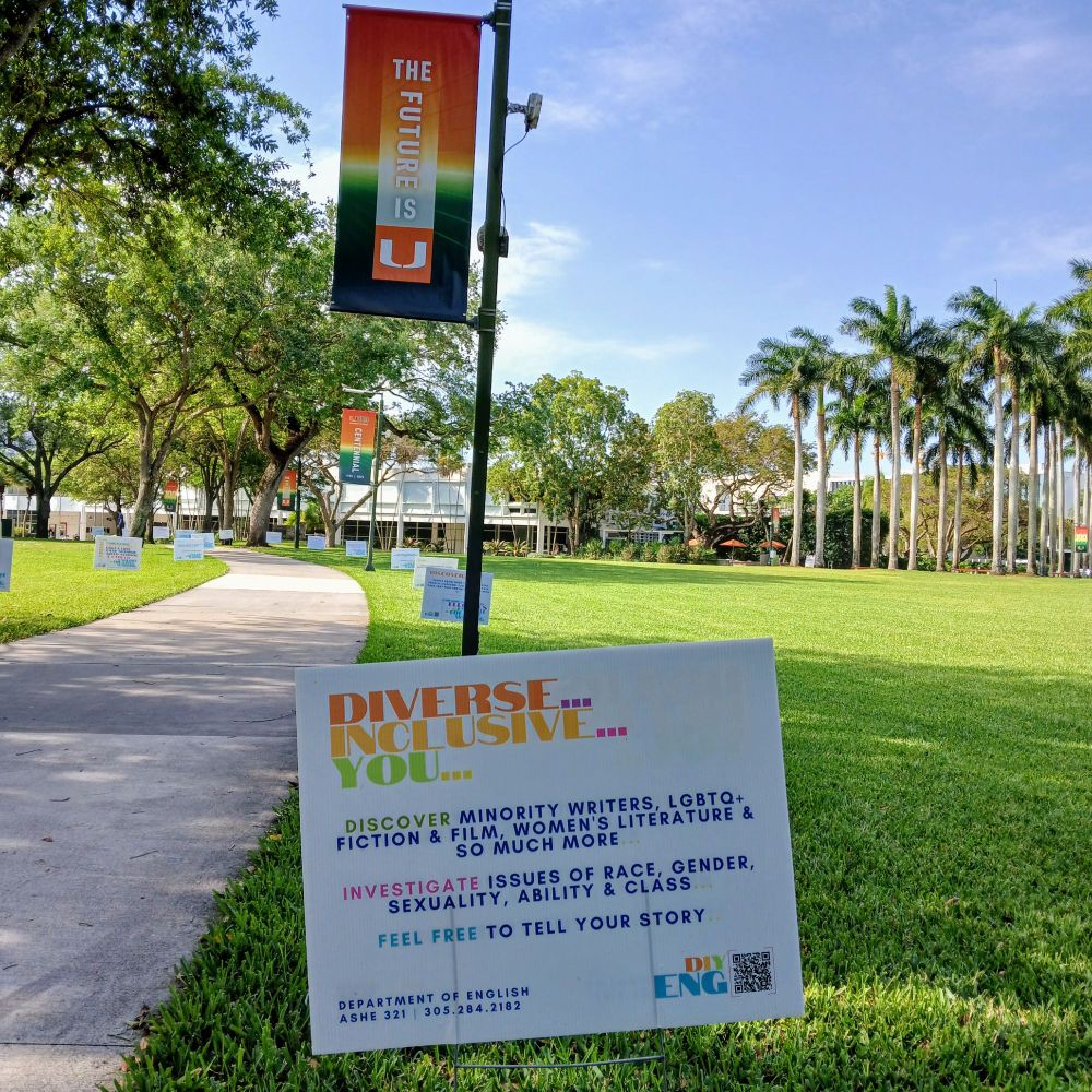 A sign promoting the English & Creative Writing Department at the University of Miami in the foreground, with grass, palm trees, and a series of banners behind. The sign reads "Diverse..Inclusive...You...Discover Minority Writers, LGBTQ+ Fiction & Film, Women's Literature, & So Much More // Investigate Issues of Race, Gender, Sexuality, Ability, & Class // Feel Free to Tell Your Story"