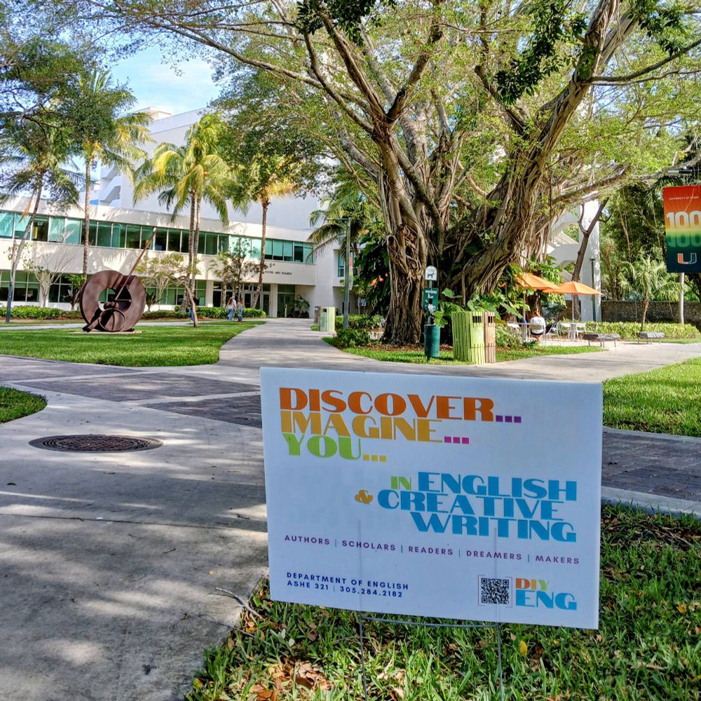 A sign promoting the English & Creative Writing Department at the University of Miami planted in a patch of grass in front of a large banyan tree, a piece of public sculpture, and the main administration building of the university that also houses the humanities departments at UM. The sign reads "Discover...Imagine...You...in English & Creative Writing / Authors / Scholars / Readers / Dreamers / Makers"