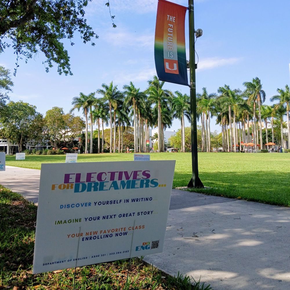 A sign promoting the English & Creative Writing Department at the University of Miami in the foreground, with grass, palm trees, and banner behind. The sign reads "Electives for Dreamers... Discover Yourself in Writing // Imagine Your Next Great Story // Your New Favorite Class Enrolling Now". The banner behind reads "The Future is U".