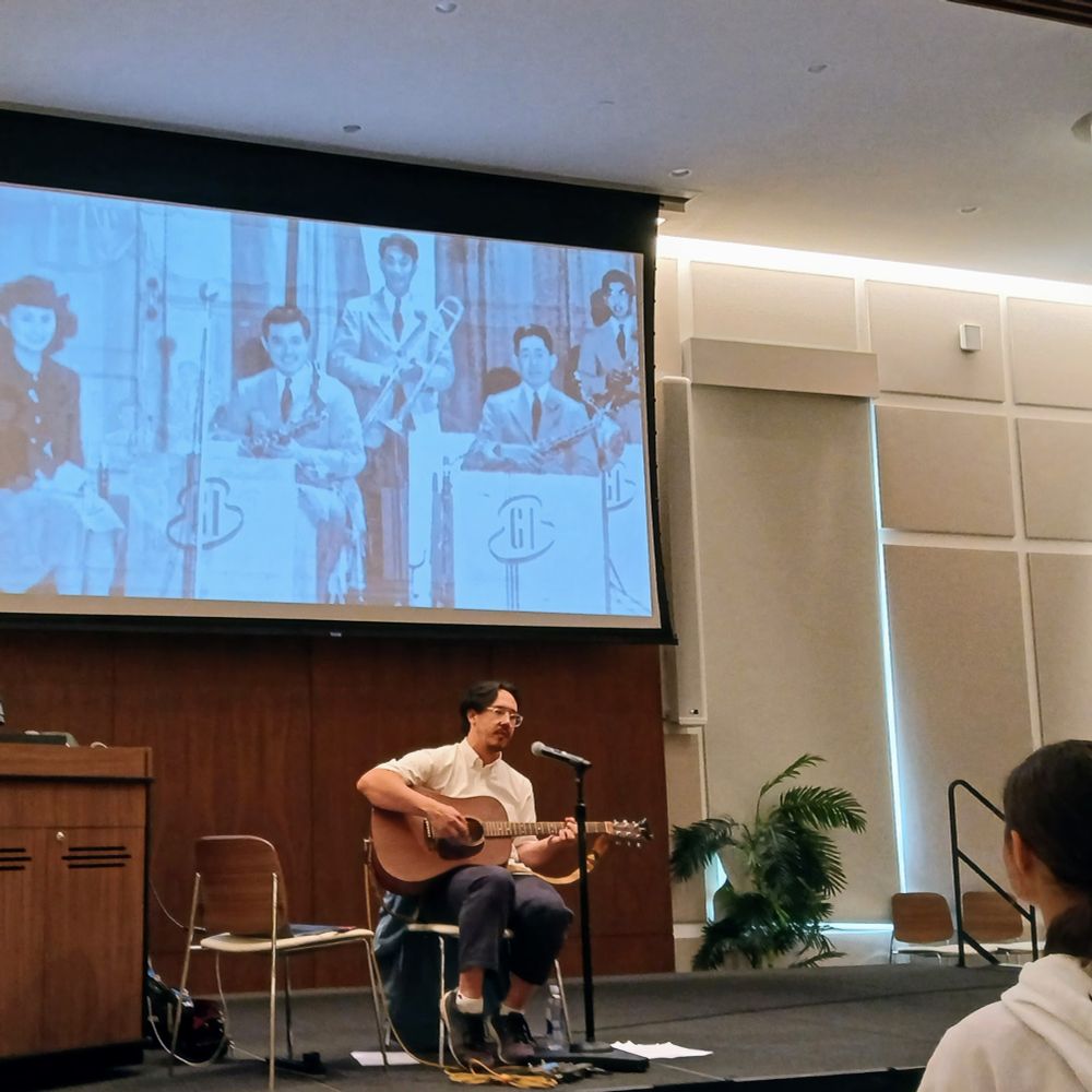 Julian Saporiti performing his song "The Best Goddamned Band in Wyoming" in the Kislak Center of University of Miami Libraries on Friday November 15, 2024, sitting on a chair strumming an acoustic guitar, with an image of the George Igawa Orchestra, a swing band formed by incarcerated Japanese Americans in Heart Mountain, Wyoming in the early 1940s, projected on a large screen behind him.