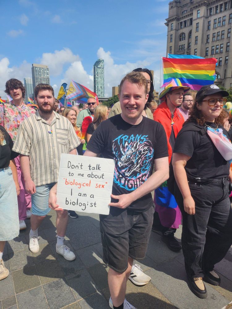 A picture of the Liverpool Pride 2025 march. The focus of the picture is a man in amongst the crowd of people marching together near the Liver building in Liverpool. The man is wearing a black tshirt with a dragon graphic on it and "trans rights" written on it. He is holding a placard that says: Don't talk to me about "biological sex" I am a biologist.