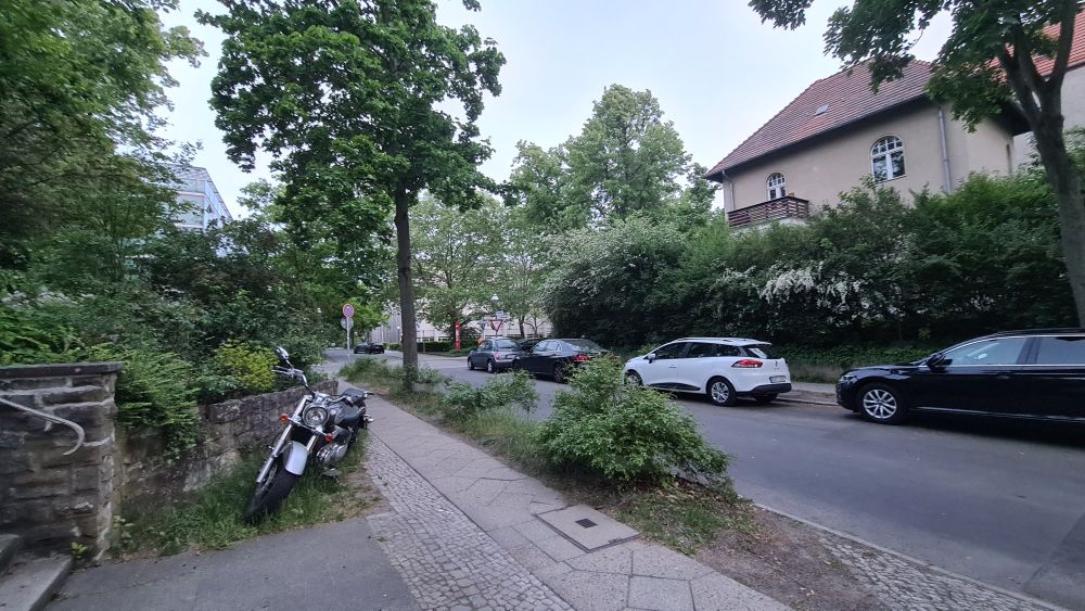 A picture of a street on Freie Universität Berlin campus. It is a spring evening, and the plant life is vibrant green and lucious. A motorbike sits on the corner of the pavement, out of the way of pedestrians. In the distance poking out between the trees there are some campus buildings, and the Seminaris conference centre.
