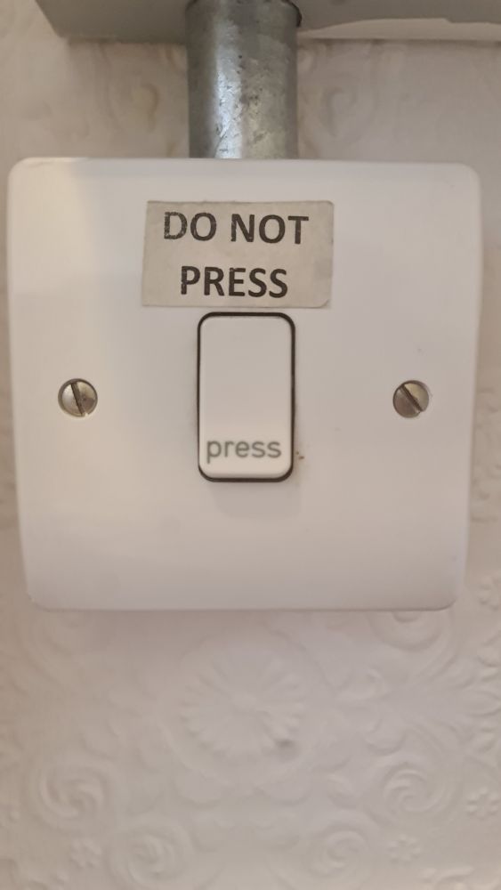 A wall mounted switch, similar in style to a light switch. The button is marked "press" but someone has attached a label above it that says "DO NOT PRESS"
