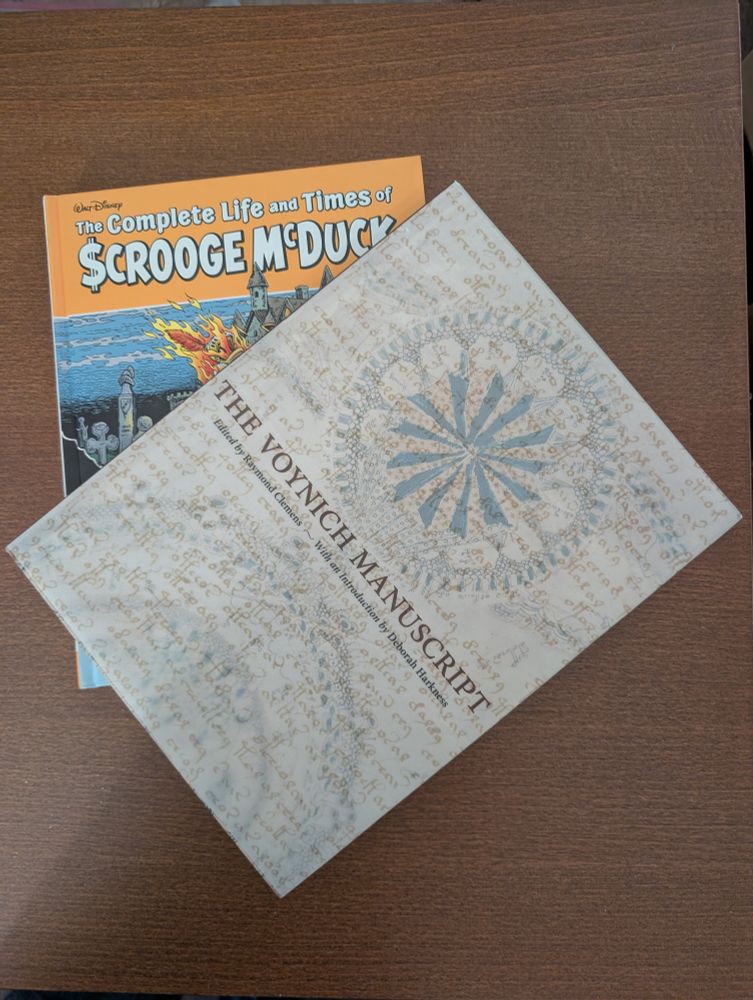 A copy of the Voynich Manuscript sitting on top of The Complete Life and Times of Scrooge McDuck volume 1.