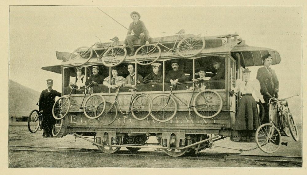 A small two-axle streetcar with bicycles hanging off the sides, ends, and roof.  Nine are visible, presumably there are more on the other side.