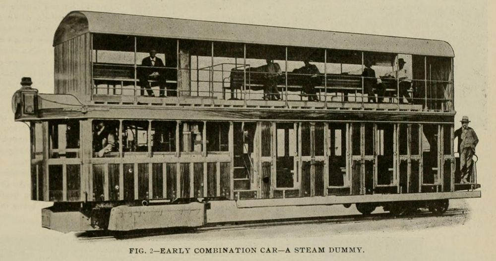 A double-decker self-propelled railroad car.  At the front is an enclosed cab/boiler room, behind that some stairs to the second level, and then four doors to enclosed cabins.  The top level has a roof but open sides and long benches for seating.  There is skirting hiding the front driving wheels and an exposed unpowered truck (bogie) in back.