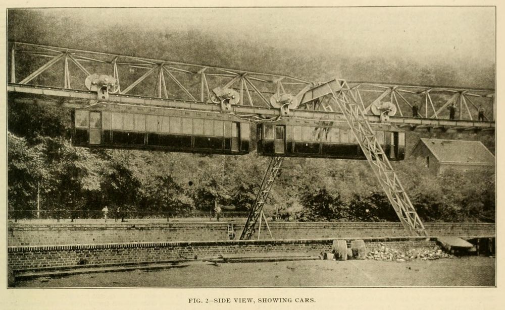 A two-track hanging monorail, suspended by spindly truss legs over a river, viewed from the side, showing two cars.