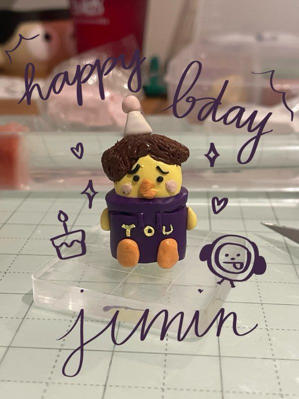 A polymer clay recreation of Jimin of BTS's birthday cake from a few years ago. It features a yellow chick wearing a birthday hat and a purple hoodie that says "YOU". Edited on the photo is writing that states "happy bday jimin" with various small drawn hearts, sparkles, and a birthday cake. 