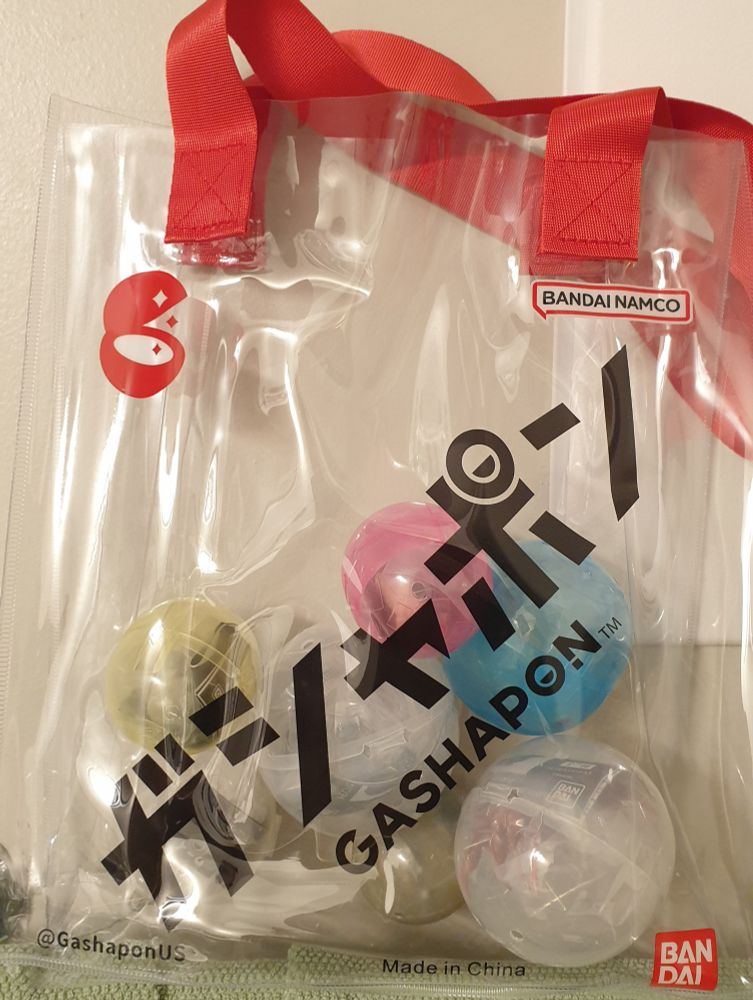 A tote bag made of thick, clear plastic material (like something to carry toiletries or beach stuff in) with red nylon handles, emblazoned with Gashapon & Bandai logos.  Several gashapon capsules in various sizes/colors are visible within 