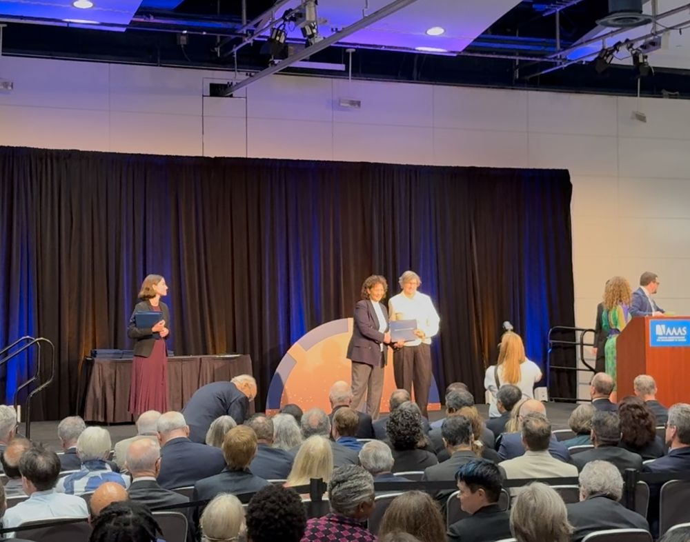 a stage with a blue-lit black curtain behind it & a crowd seated facing it. a woman in a maroon dress stands to the left holding a stack of awards. Two women stand center stage. Ine in a brown blazer is presenting Kate Aubrecht, in a white jacket, with her AAAS Fellow plaque. 
