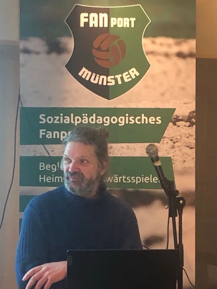 Andreas Kemper sitzt beim Vortrag des Fanprojektes FANport Münster vor einem FANport-Logo