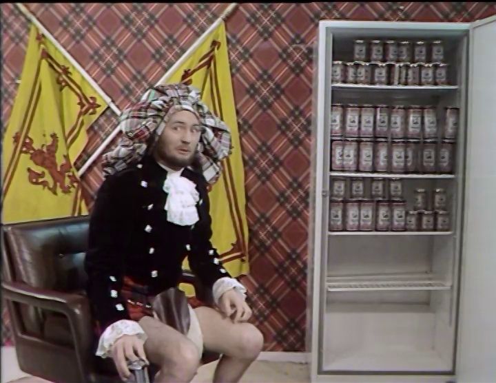 Kenny Everett dressed in a ridiculous Scottish outfit with oversized bunnet and undersized kilt, sitting in front of a fridge full of Younger's Tartan cans