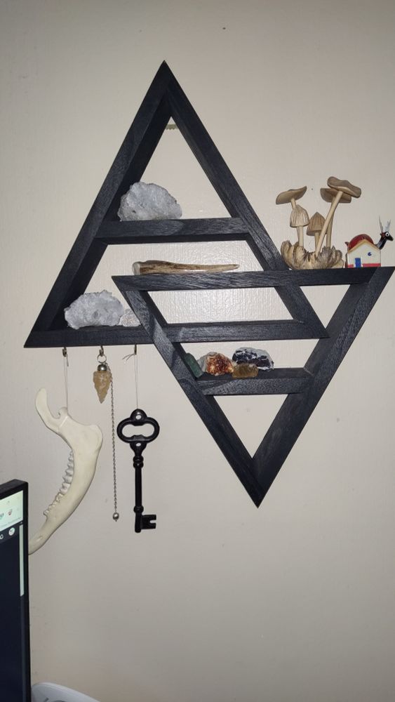 A black painted wooden shelf in the shape of two intersecting triangle. On the many small shelves sit: various crystals (a split geode on two different levels, some desert rose, citrine in two different clusters, aventurine, and a peacock ore), a wooden mushroom carving, a tiny ceramic house charm, an antler whistle, and a bobble headed alebrije. Hanging from a trio of hooks are: a big metal skeleton key, a citrine pendulum, and a deer jawbone.